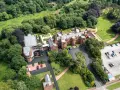 Aerial view of the abbey guest house, a grand brick building surrounded by lush green trees and manicured lawns. Cars are parked in a lot.