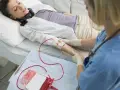 Kobieta leży w łóżku szpitalnym, słuchając muzyki przez słuchawki. Pielęgniarka wykonuje przetaczanie krwi, podłączając worek z krwią do jej ręki.