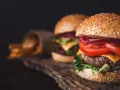 Dwa soczyste burgery z serem, pomidorem i czerwoną cebulą na drewnianej desce, obok frytki.