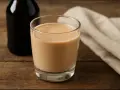 Szklanka kremowego likieru, idealny likier Baileys zamiennik, obok ciemnej butelki i lnianej serwetki na drewnianym stole.