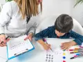 Bambino impegnato in un test per l'autismo, colora forme geometriche guidato da un'insegnante.
