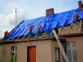 Robotnicy na dachu kryjącym niebieską plandeką. Zastanawiasz się, ile kosztuje pozwolenie na budowę?