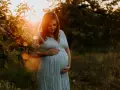 Piękne zdjęcia w ciąży. Kobieta w błękitnej sukience trzyma dłonie na brzuchu, otoczona ciepłym światłem zachodzącego słońca.