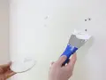 Persona aplica masilla en una pared blanca con una espátula azul para reparar agujeros.