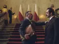 Hubert Niedziela, w garniturze, trzyma teczkę, rozmawiając z mężczyzną na schodach przed polskimi flagami.