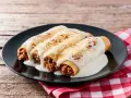 Canelones de carne picada cubiertos de queso fundido, servidos en un plato negro sobre una mesa de madera rústica.