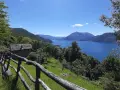 Sentiero del Viandante tappe: un casolare in pietra con tetto in ardesia e un recinto in legno rustico si affacciano su un lago blu intenso circondato da montagne verdi.