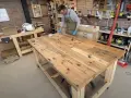 Uomo con grembiule lavora su un banco da lavoro con pallet, applicando finitura al legno. L'officina è piena di attrezzi e materiali.
