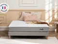 Matelas gris et blanc avec logo "mello", sur un sommier, avec des coussins et un plaid.