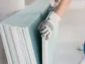 Ręka w rękawicy chwyta stos płyt regipsowych, przygotowując się do remontu.