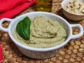 Pesto di melanzane cremoso in una ciotola bianca, guarnito con una foglia di basilico. Sullo sfondo, mandorle e olio d'oliva.