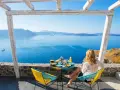 Mujer desayunando en balcón con vista al mar azul y montañas.