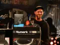 Un DJ souriant pose avec des prix et du matériel, dont des casques Sennheiser et un tourne-disque Numark. Une liste DJ internationale est visible.