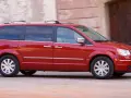 Czerwony Chrysler Grand Voyager w ruchu, z przyciemnianymi szybami i relingami dachowymi, przemierza ulicę obok kamiennej fasady.