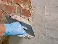 Hand mit blauer Handschuh glättet mit Kelle den **Sanierputz Keller** auf rotem Ziegelmauerwerk.