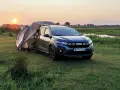 Dacia Jogger Camper rozłożony na biwaku o zachodzie słońca. Namiot połączony z autem, otoczony łąką i wodą.