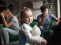 Smutna dziewczynka z misiem w objęciach, podczas gdy rodzice w tle przeżywają trudne chwile. Temat: rozwód a dzieci.