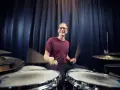 Ein lächelnder einarmiger Schlagzeuger mit Brille spielt begeistert Schlagzeug vor einem dunklen Vorhang.