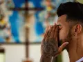 A man with tattoos prays before a crucifix, contemplating if is vaping a sin.