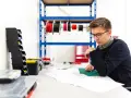 Młody mężczyzna w okularach analizuje dokumentację techniczną. Na stole leżą narzędzia, a w tle widać półkę z przewodami, co sugeruje pracę nad projektem z dziedziny plan bioz.