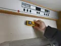 Hand håller fuktmätare mot vägg för fukttest protokoll.