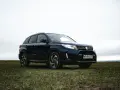 Ciemnoniebieski Suzuki Vitara Hybrid na tle zachmurzonego nieba, zaparkowany na trawiastym wzgórzu.