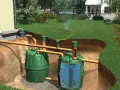 Regenwasser wird gesammelt und im Garten zur Bewässerung genutzt. Zwei grüne Tanks speichern das Wasser, das über Rohre vom Haus und aus dem Garten zugeführt wird.