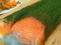 Un gran filete de salmón marinado casero cubierto de eneldo fresco. En el fondo, finas lonchas de salmón ya preparadas.