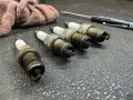 Four used spark plugs lie on a textured surface next to a rag and a screwdriver. This image might make you wonder how many spark plugs a car has.