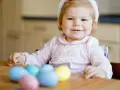 Kleines Mädchen mit Hasenohren und bunten Ostereiern. Sie sitzt am Tisch und schaut neugierig auf das **Baby Ei**.
