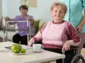 Starsza kobieta w wózku inwalidzkim, ocena jej sprawności według skali Barthel. W tle druga seniorka przy balkoniku.