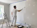 Mężczyzna w białym ubraniu maluje ścianę wałkiem. Na podłodze folia ochronna, drabina i kuweta z farbą. To pierwszy grunt.