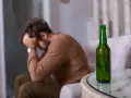 Mężczyzna siedzi z głową w dłoniach, obok stoi zielona butelka. Czuje duszności po alkoholu.