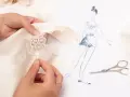 Main d'une couturière réalisant un assemblage couture délicat sur un tissu de soie, avec un croquis de mode et des ciseaux à proximité.