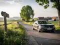 En mörkgrå bil kör på en slingrande väg genom ett lantligt europeiskt landskap med fält och hus.