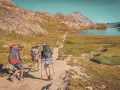 Trois randonneurs équipés pour le tour du Queyras traversent un sentier de montagne, un lac bleu et des sommets rocheux en toile de fond.