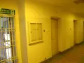 Korytarz więzienny z kratami i drzwiami cel. Nad drzwiami znak "Wyjście ewakuacyjne".