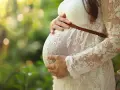 Brzuch kobiety w ciąży, otulony koronkową sukienką. Delikatne światło i zieleń natury tworzą ciepłą atmosferę, jakby wspierały jej luteina dopochwowa w ciąży.