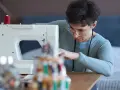 Kobieta w okularach skupiona na szyciu lamówki na maszynie. W tle widać stojak z kolorowymi nićmi.