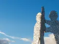 Der Molecule Man in Berlin, eine Skulptur aus Metall mit vielen Löchern, ragt gegen einen blauen Himmel mit Wolken.