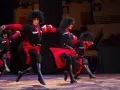 Tancerze baletu gruzińskiego Sukhishvili w tradycyjnych strojach z czarnymi czubami i czerwonymi akcentami wykonują dynamiczny taniec na scenie.