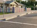 Parking, chodnik i kolorowy płotek oddzielają plac zabaw od miejsc parkingowych. Odległość miejsc parkingowych od granicy działki jest widoczna.