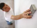 Un homme ponce un mur blanc avec une cale à poncer, préparant la surface avant la peinture.