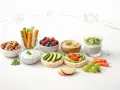 Zdrowe przekąski: warzywa pokrojone w słupki, owoce jagodowe, hummus, ryżowe wafle z awokado i pomidorkami, deser chia z kiwi, orzechy i jogurt.