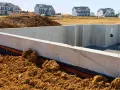 Betonowa ława fundamentowa w wykopie, otoczona ziemią. W tle nowe domy w budowie.