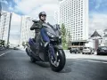 Mężczyzna w kasku i okularach przeciwsłonecznych jedzie niebieskim skuterem Yamaha X-MAX 400 po miejskiej drodze. W tle wysokie budynki i samochody.
