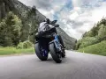 Motocyklista na Yamaha Niken pokonuje zakręt na górskiej drodze, otoczony zielenią i chmurami.