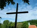 Ein großes Holzkreuz ragt vor blauem Himmel mit Wolken auf. Im Hintergrund eine Kirche und grüne Bäume. Wann ist Christi Himmelfahrt?