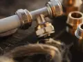 Metalowe złączki hydrauliczne, w tym trójnik i nypel, leżą na drewnianym blacie obok pęczka lnu.