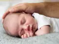 Ciepła dłoń ojca głaszcze śpiące, **2 miesięczne dziecko**. Maleństwo wtulone w miękki kocyk, wygląda na spokojne i bezpieczne.